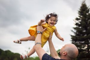 A child wearing a yellow jacket is lifted high into the air against a cloudy sky and evergreen trees.