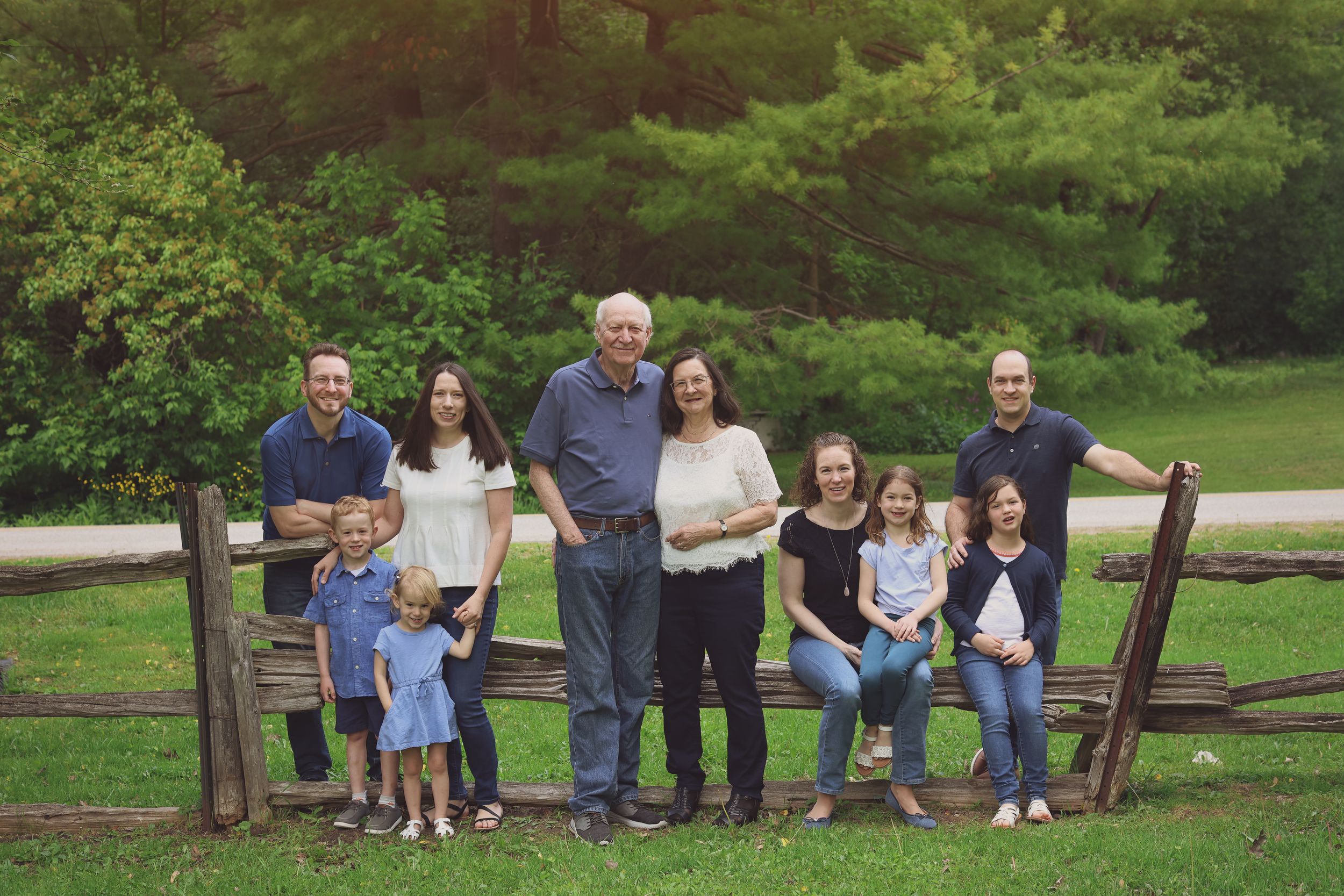 Multi-generational family portrait outdoors near rustic wooden fence surrounded by lush green trees.