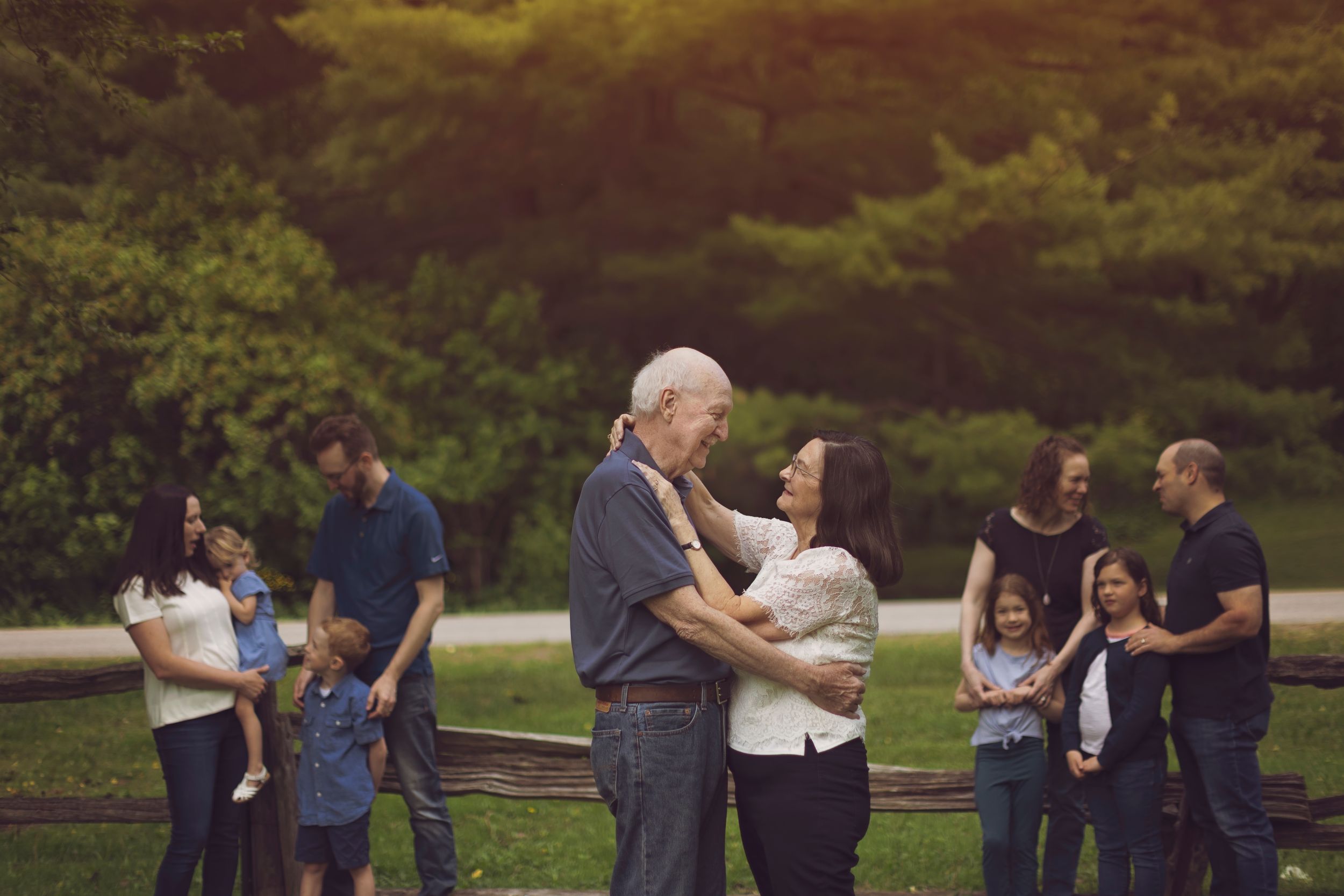 An elderly man and woman embrace outdoors during a family gathering near a wooden fence with lush green trees.