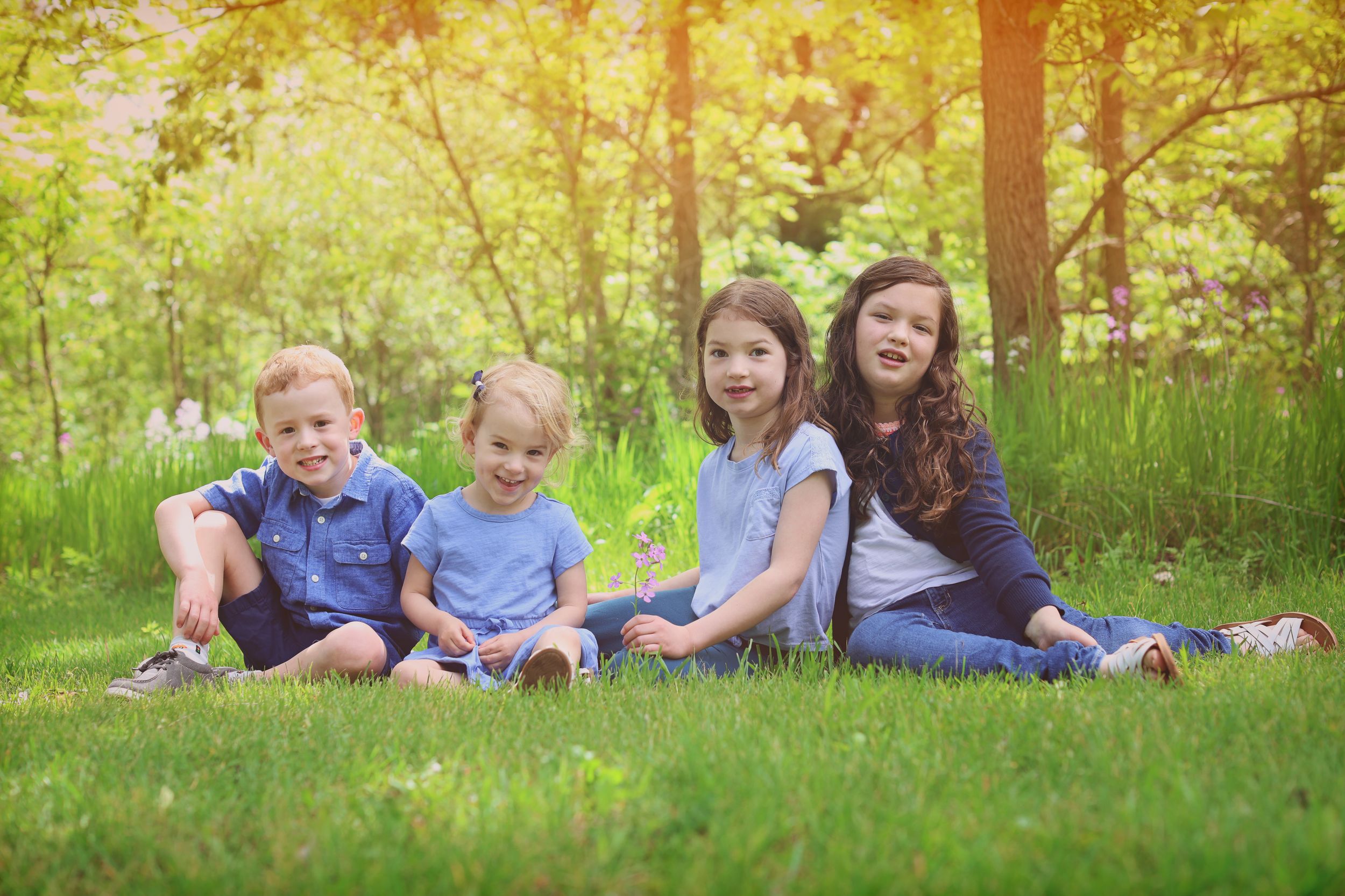 Four happy children sitting together on green grass in a sunny park surrounded by lush trees.