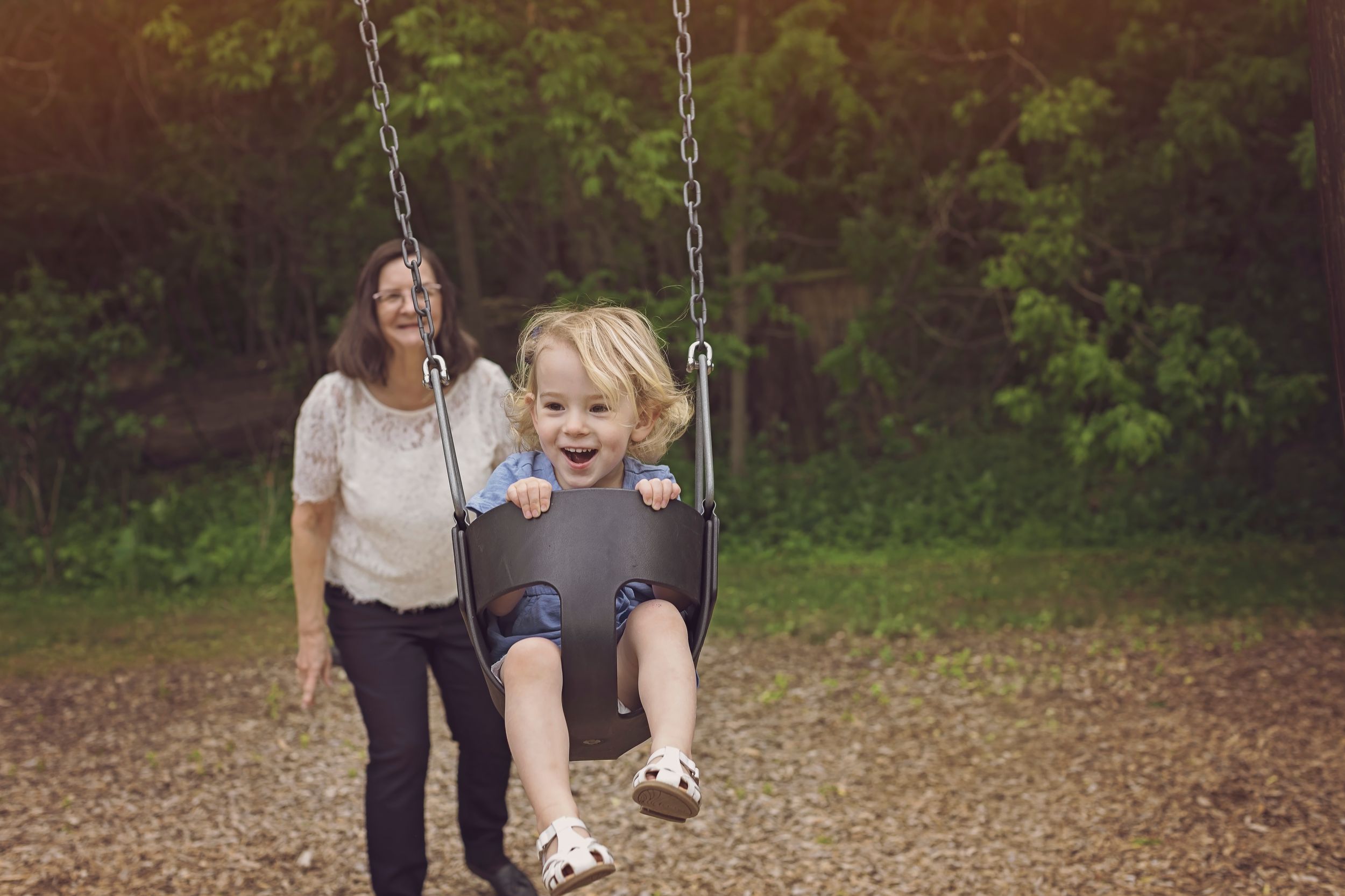 A happy child laughing on a swing being pushed by a smiling woman in a sunny outdoor park setting.