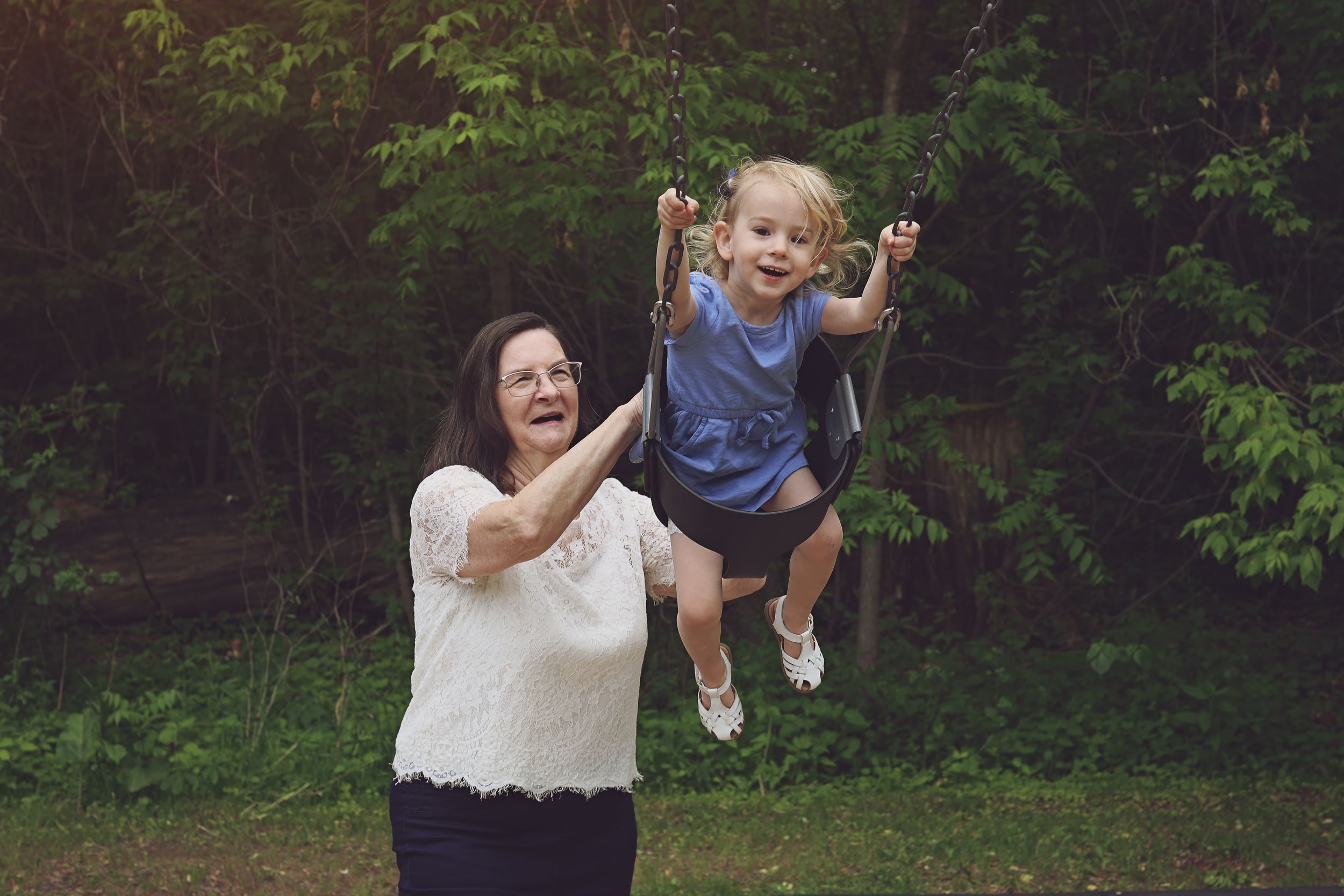 Grandmother joyfully pushes young toddler on a swing outdoors, surrounded by lush green trees.