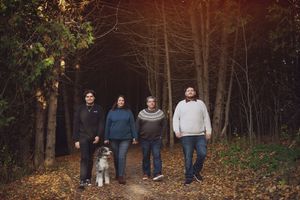 A group of people stand together in a wooded area with fallen autumn leaves on the ground during golden hour.