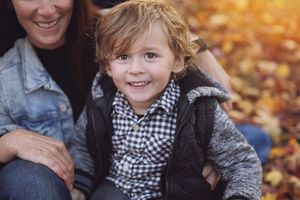 A child wearing a black and white checkered scarf smiles warmly while sitting among autumn leaves in a park.