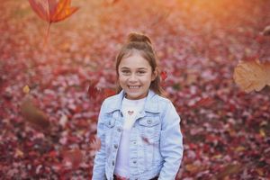 Young person in denim jacket sitting among scattered red autumn leaves.