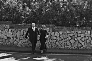 Two figures walking together along a stone wall on a sunny day, captured in black and white.