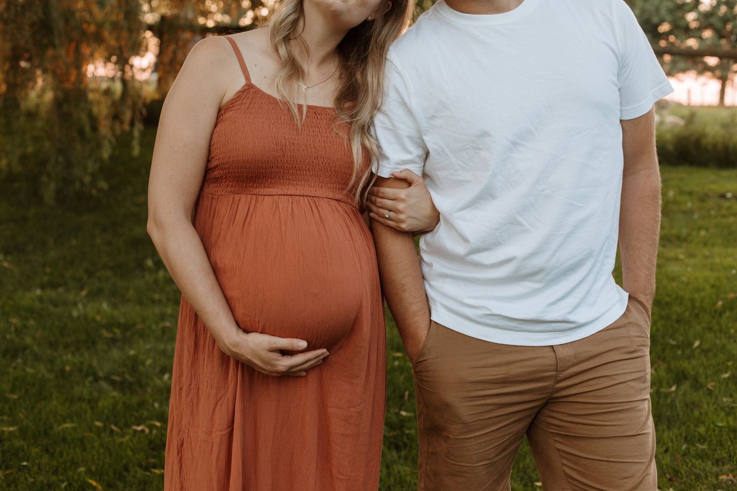 A couple embraces outdoors, with the woman wearing a rust-colored maternity dress.