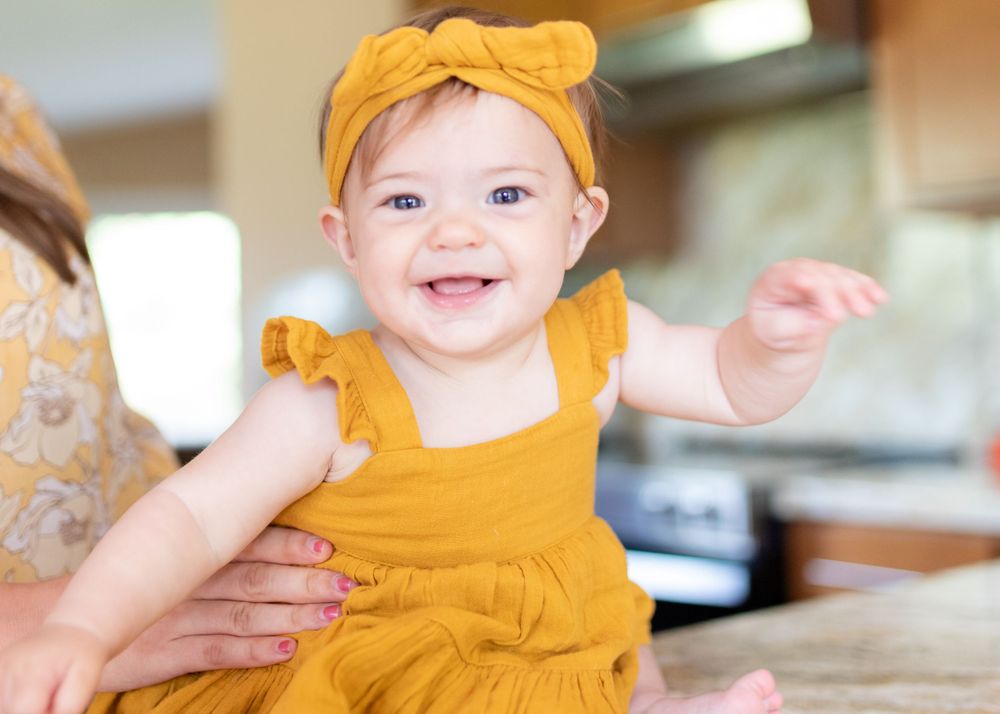 A baby wearing a yellow outfit and headband smiles brightly in a kitchen setting.