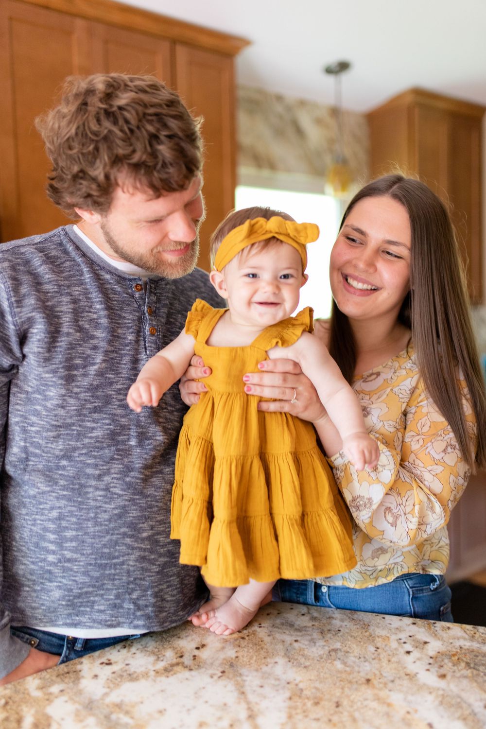 A family shares a joyful moment together in their kitchen while holding a baby wearing a mustard yellow dress.