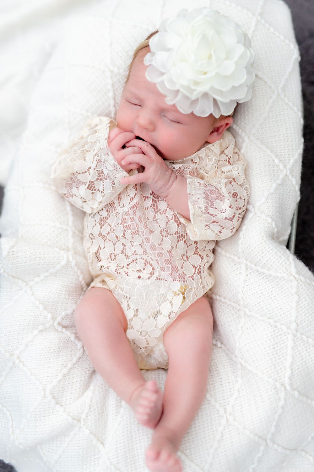 Sleeping newborn wearing white lace romper and floral headband on white blanket.