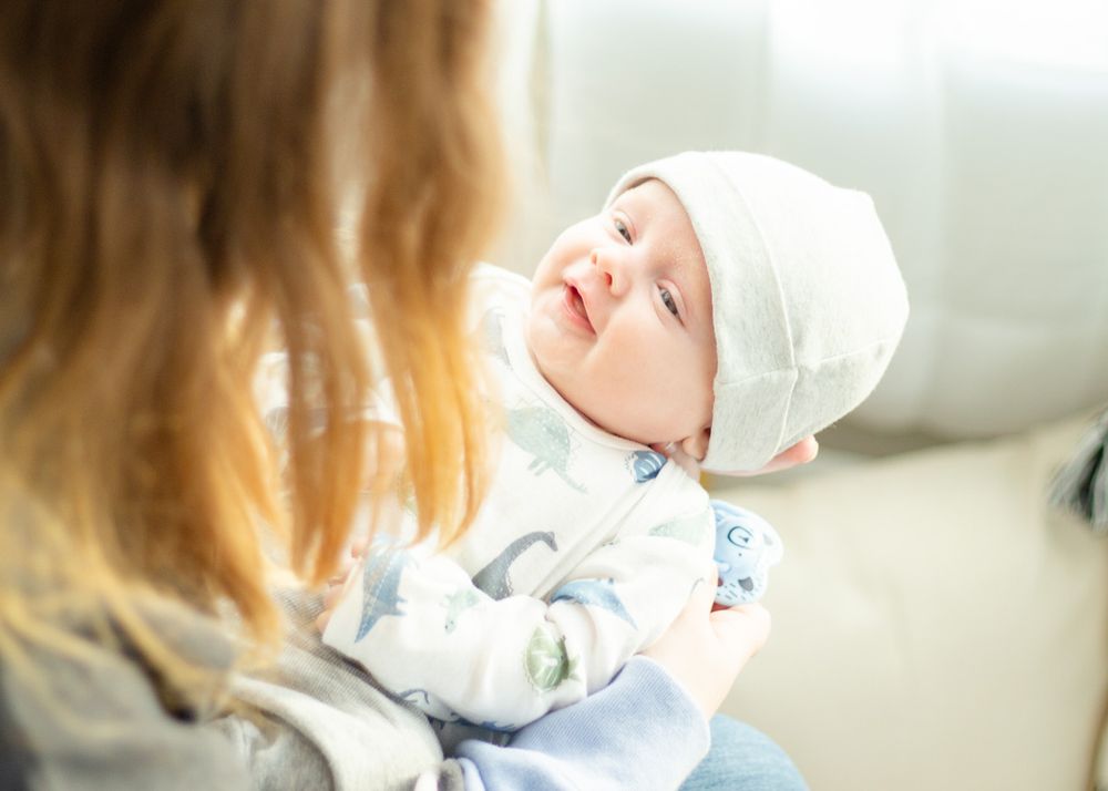 A newborn baby in a white cap smiles while being held.