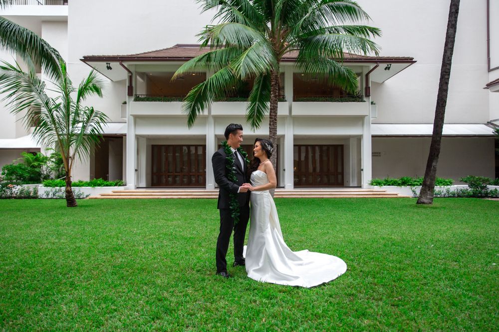 A couple shares an intimate moment on a lush green courtyard with palm trees and Halekulani Hotel Garden in the background.