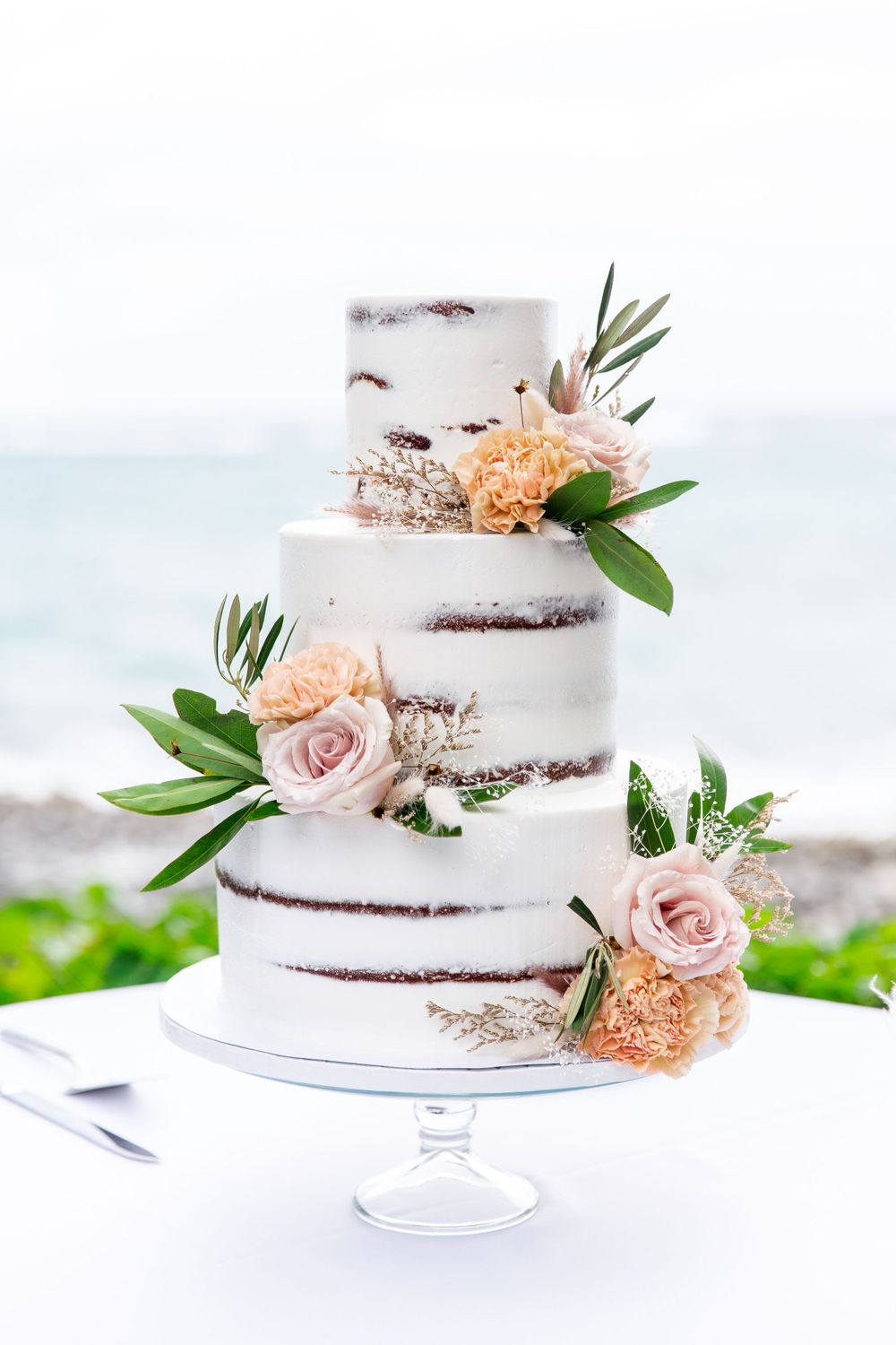 Elegant three-tier semi-naked wedding cake decorated with peach and pink roses, greenery and rustic floral accents.