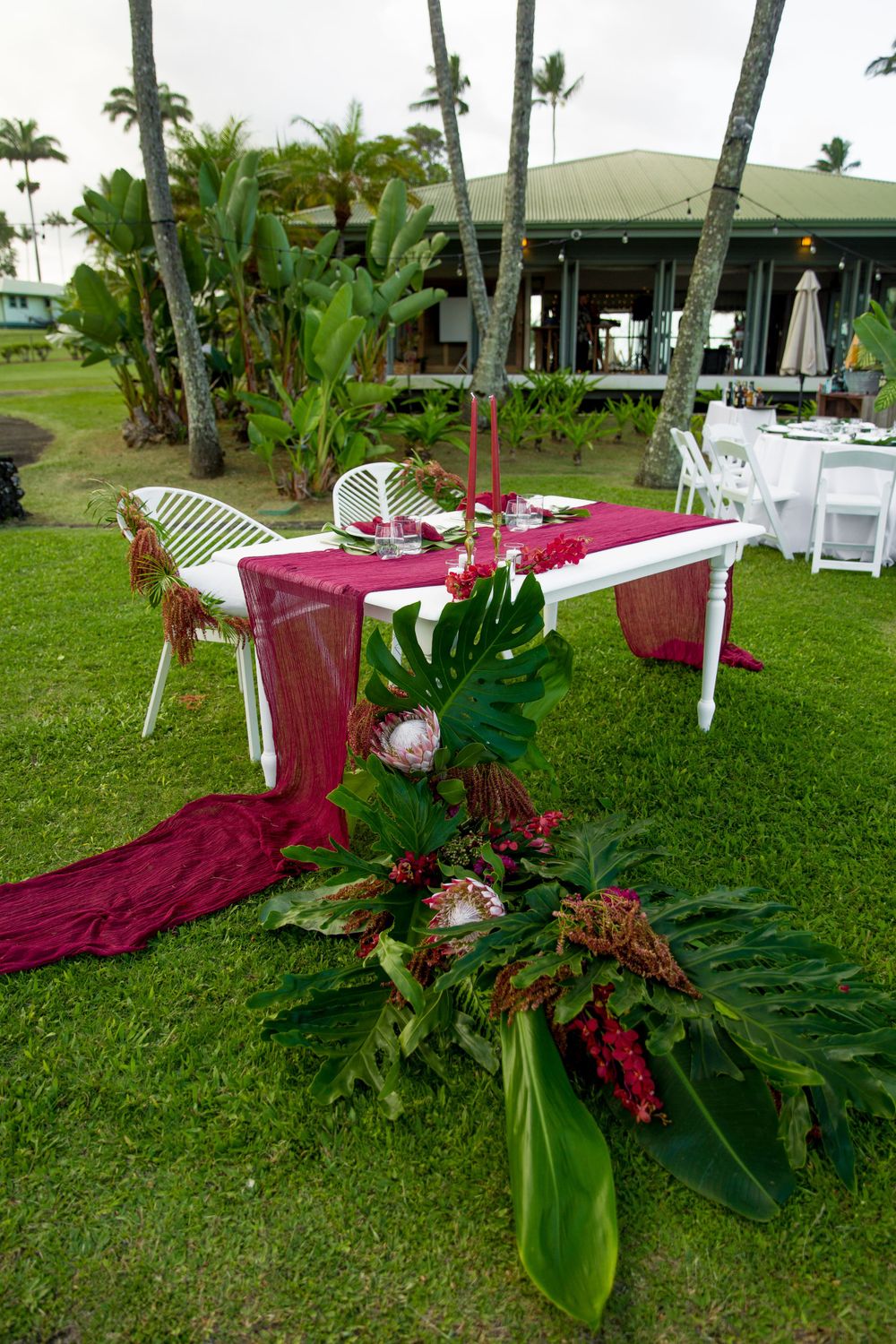 Tropical wedding table setup with burgundy runner and lush monstera leaves on grass lawn with palm trees and wedding venue behind.