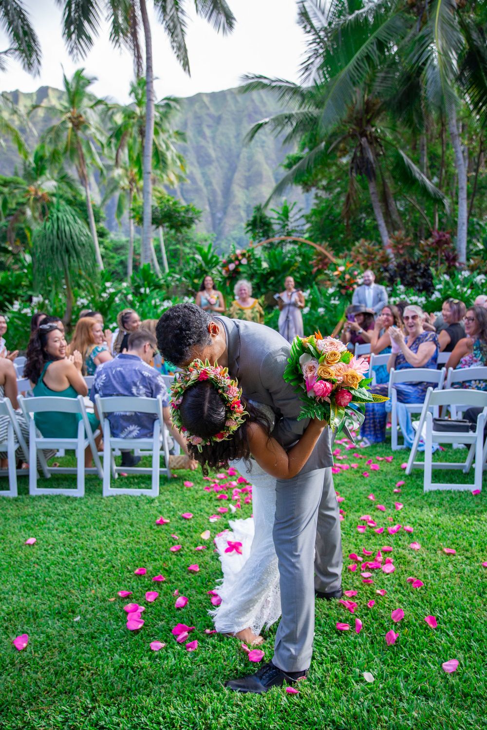 A romantic outdoor wedding ceremony in Waimanalo with palm trees, mountains, and pink flower petals scattered on green grass of Open Palms.