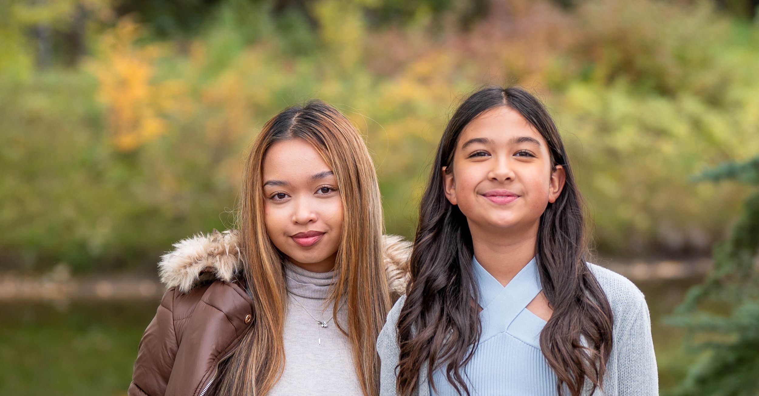 Two sisters smile softly at the camera. They are standing in a beautifal fall park with a pond behind them.