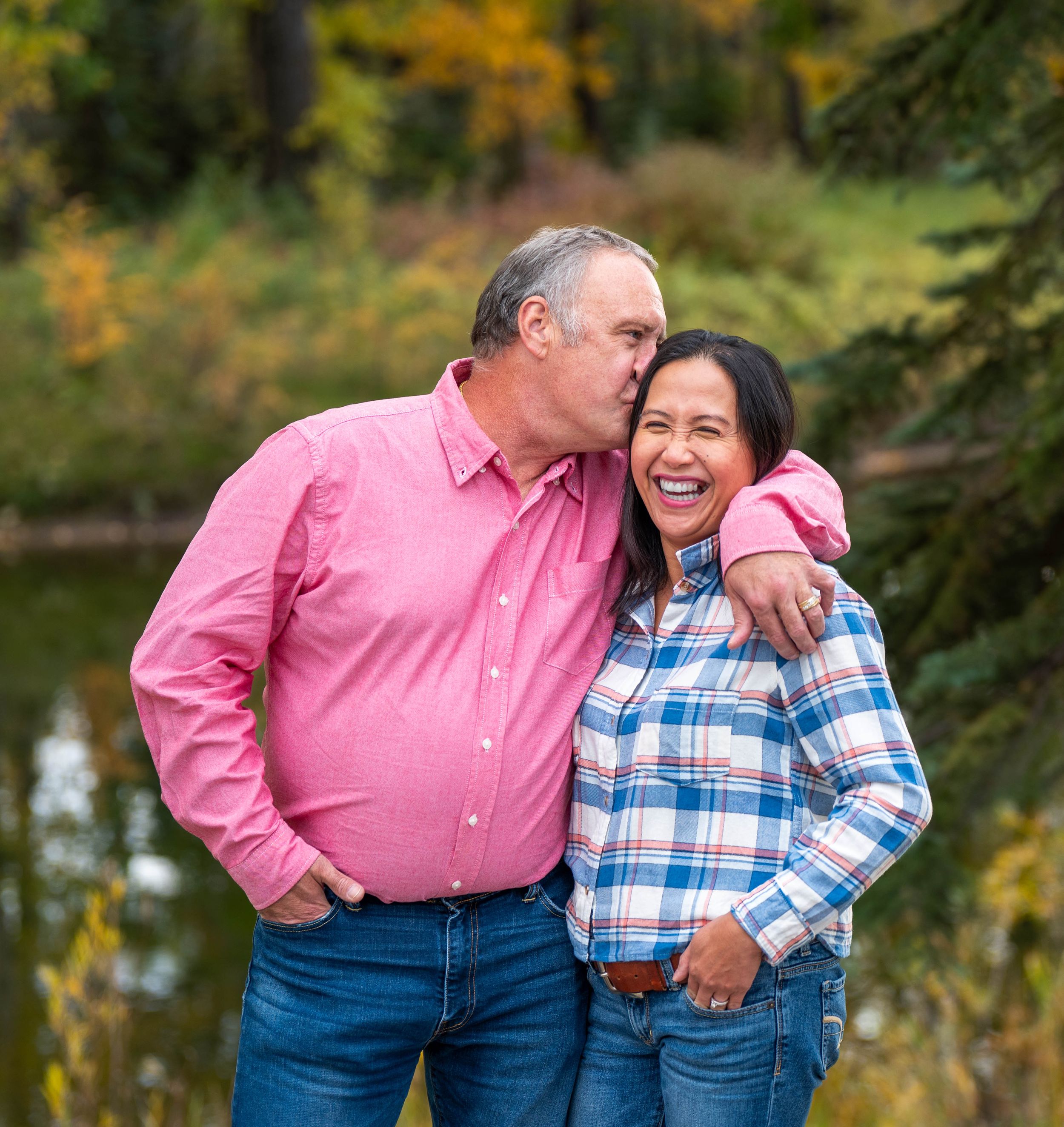A man kisses his wife on the temple as she beams. They are standing in a beautifal fall park with a pond behind them.