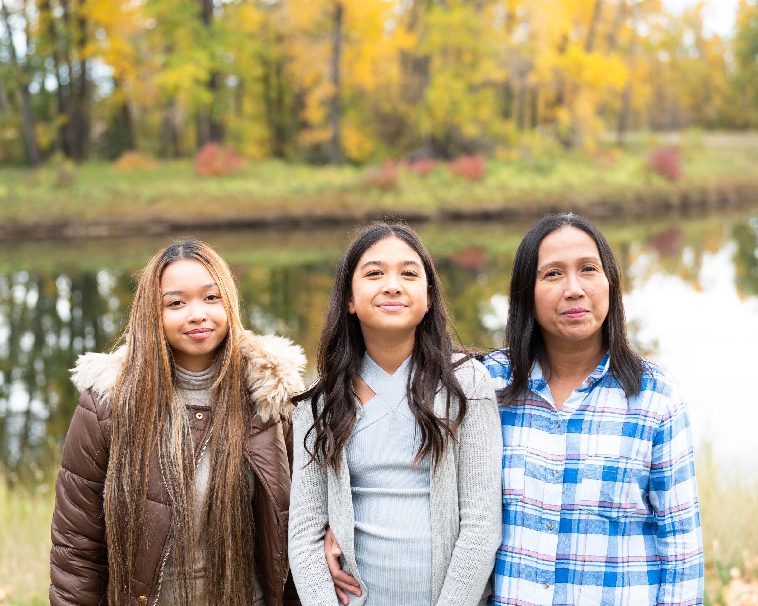 Two sisters and their mother smile softly at the camera. They are standing in a beautifal fall park with a pond behind them.