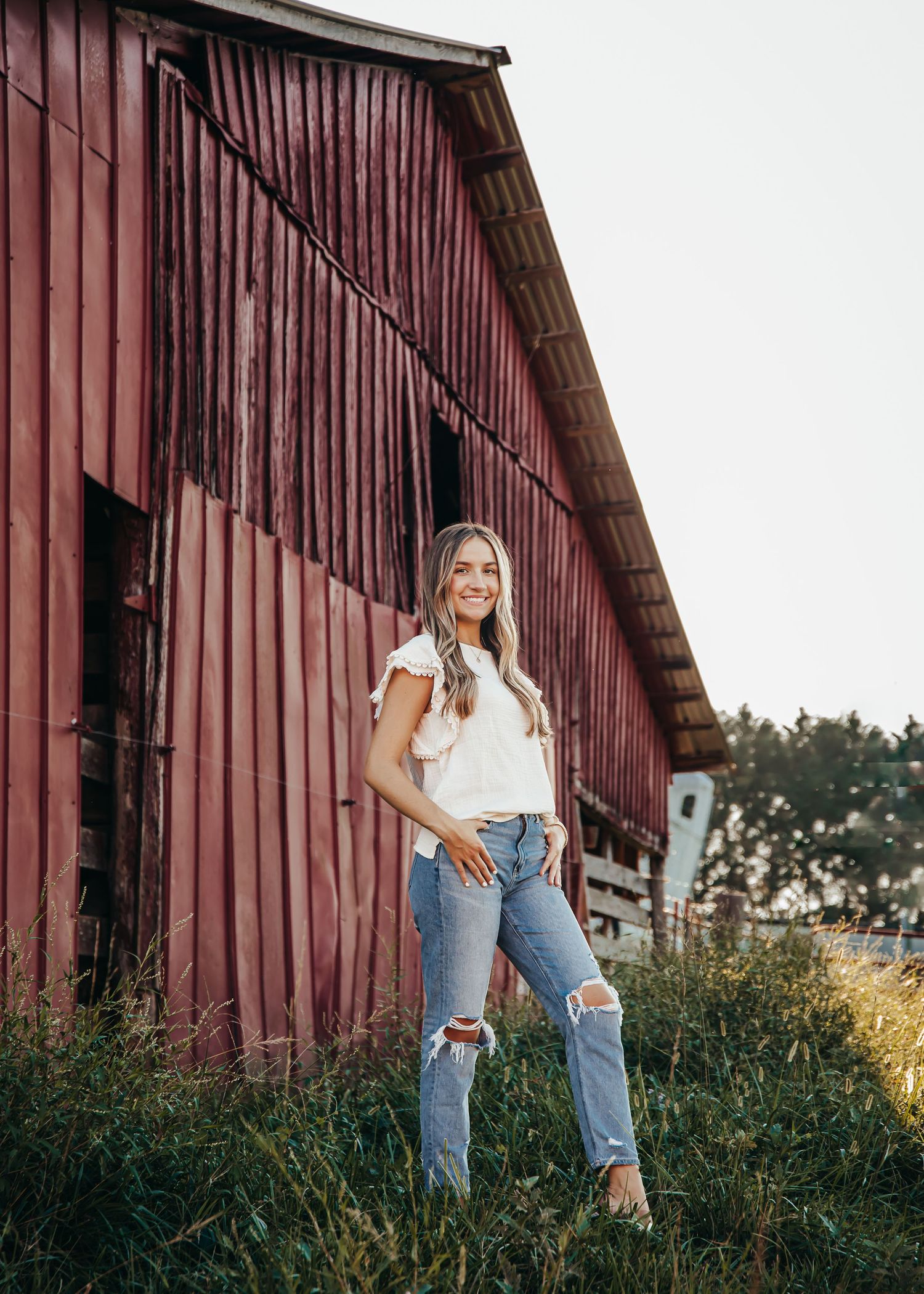 A person poses casually in ripped jeans against rustic red barn backdrop.