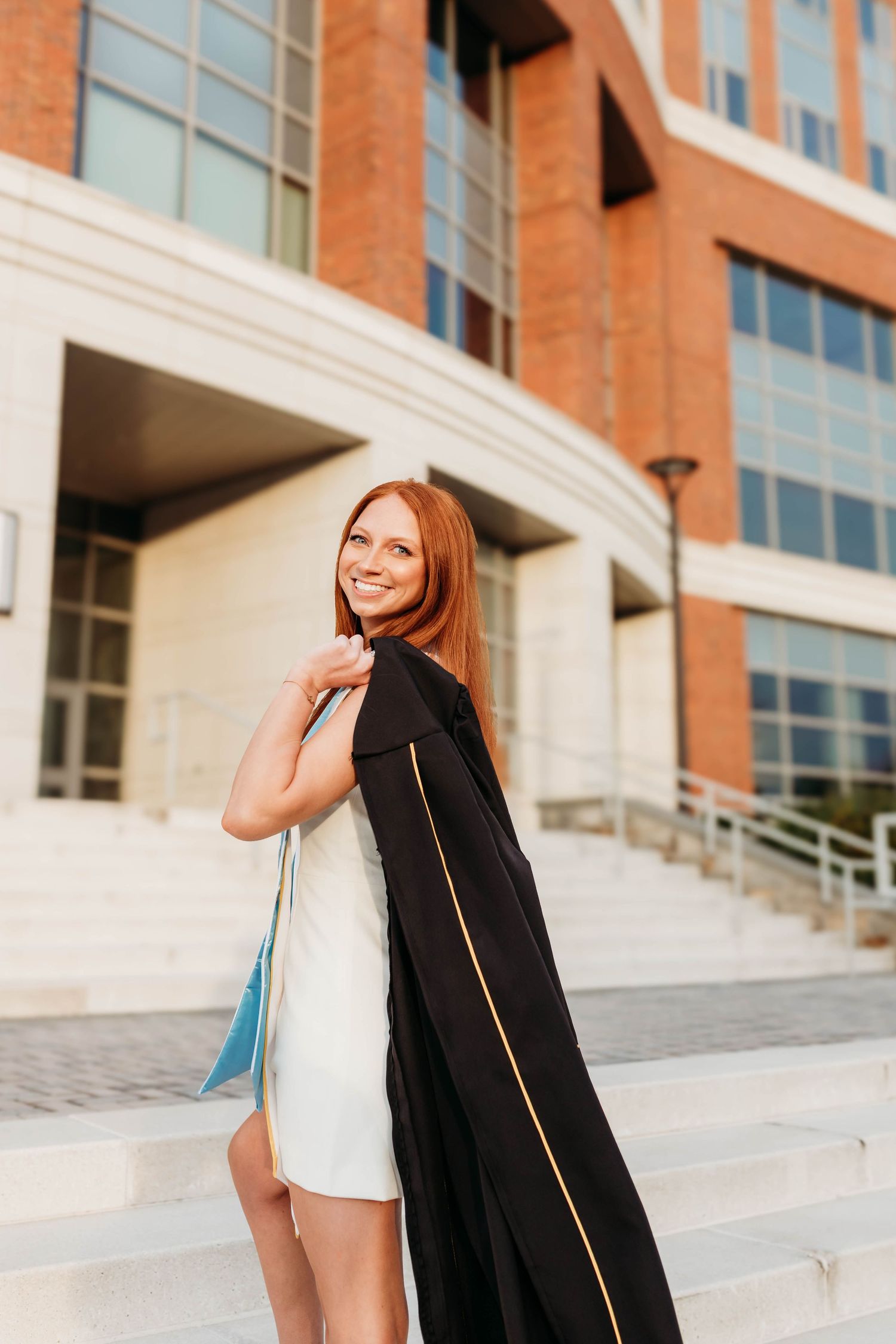 Graduate in white dress and black graduation gown poses on concrete steps of brick building.