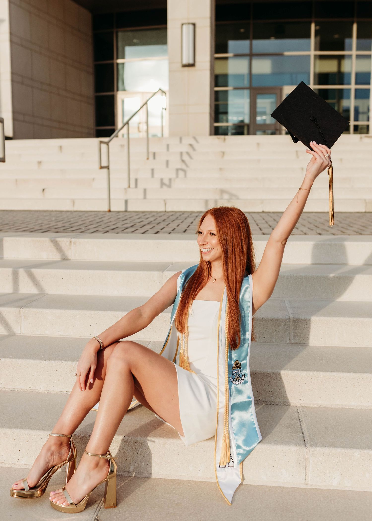Graduate sits on concrete steps wearing white dress and holding graduation cap.