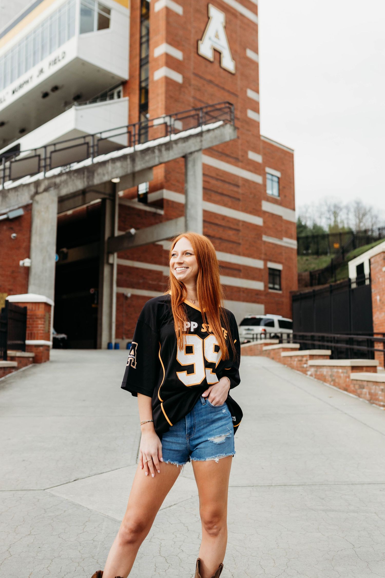 Student wearing black jacket and denim shorts poses near brick university building.