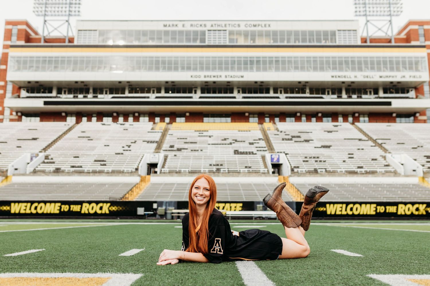 Graduate lying on football field with stadium seating visible in background.