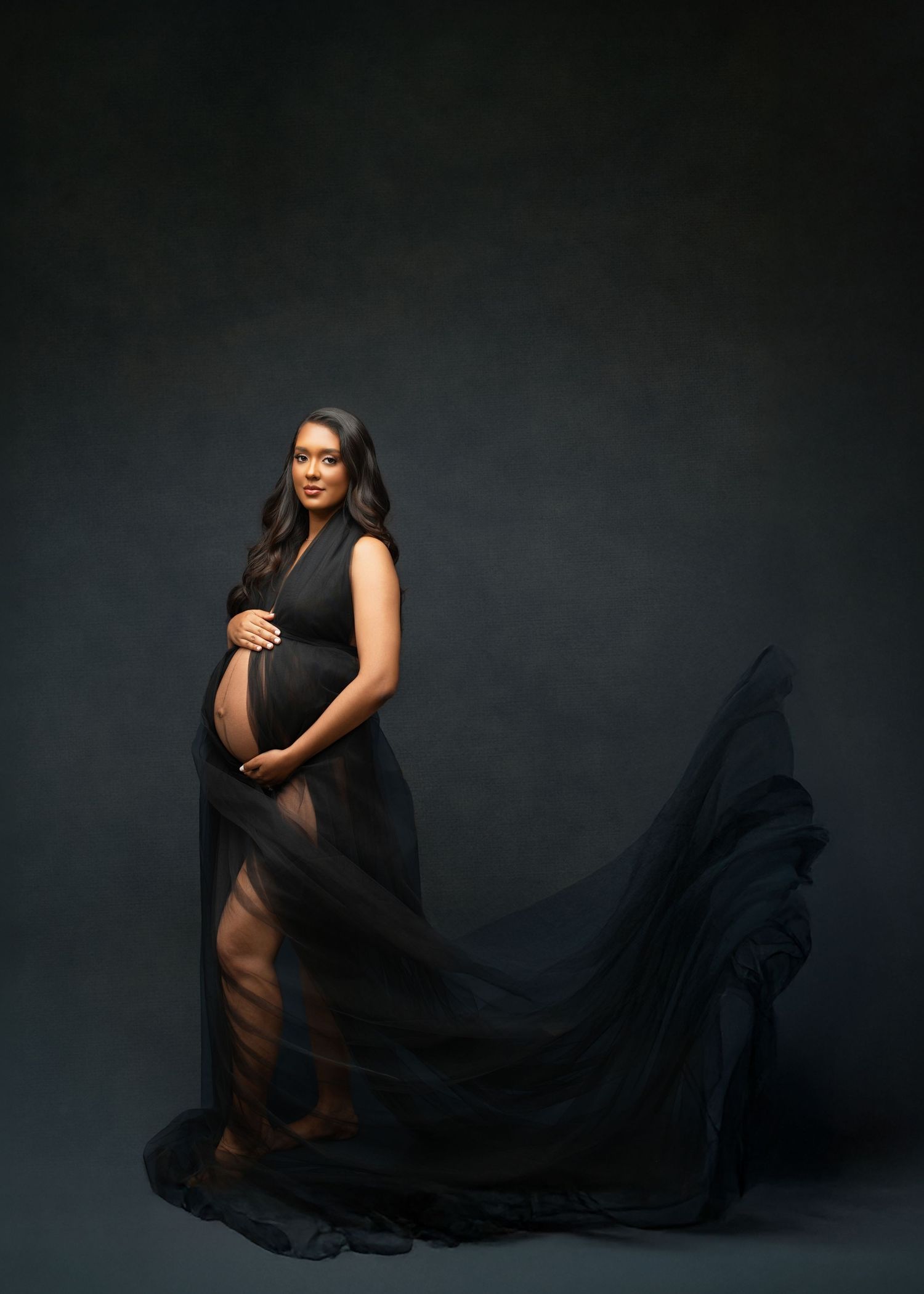 An intimate maternity portrait shows a couple embracing against a brown background, with her in a white dress and him in pink.