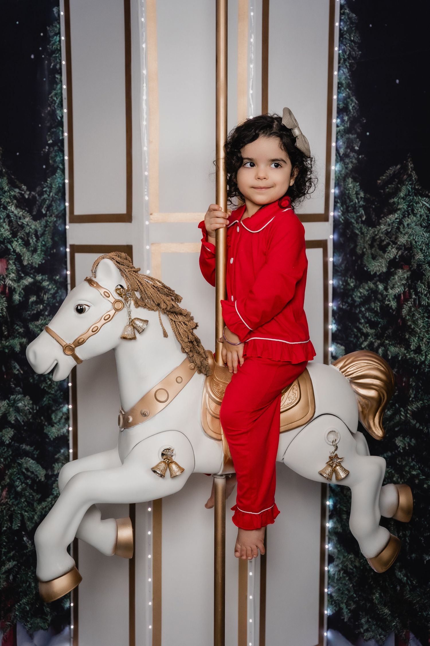 Child in red pajamas sitting on decorative white carousel horse against holiday background.