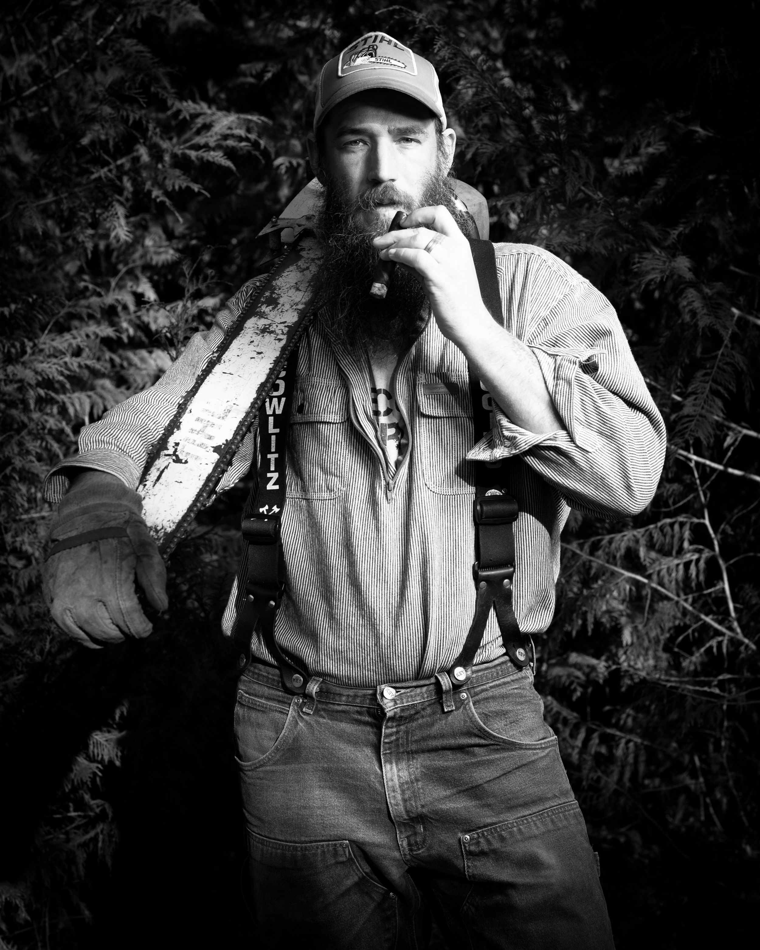 Black and white portrait photo of someone in work wear with suspenders and a baseball cap holding a chainsaw and cigar outdoors.