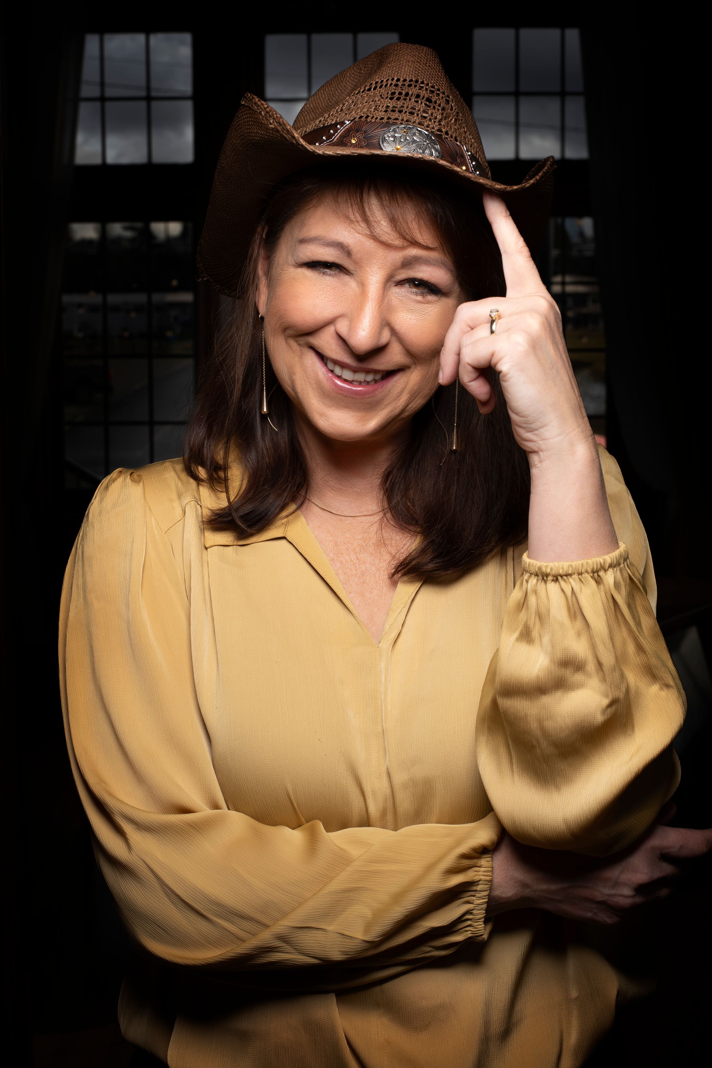 Woman in yellow blouse and cowboy hat smiling warmly against dark background.