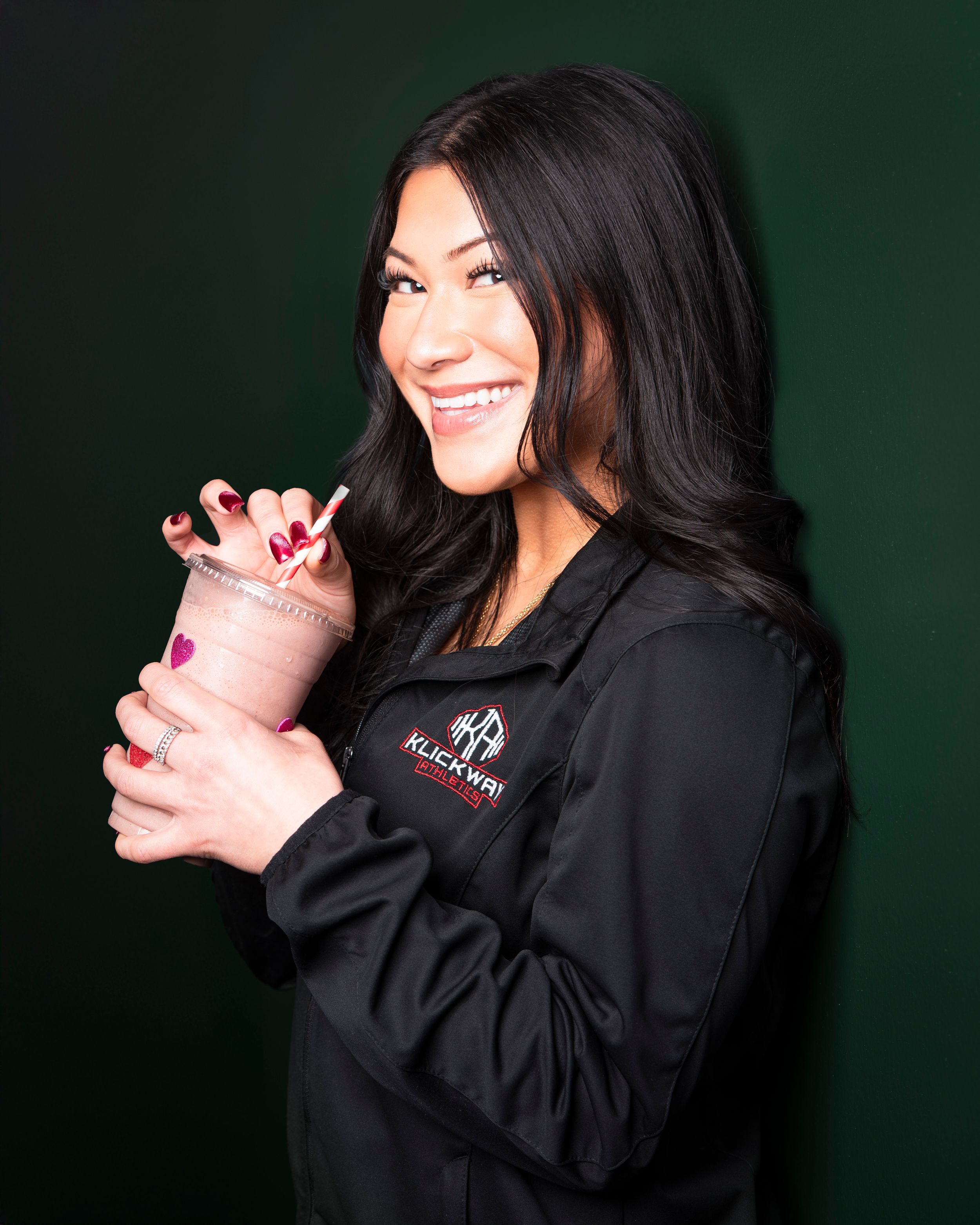 Smiling employee in black uniform jacket holding smoothie against dark green background.