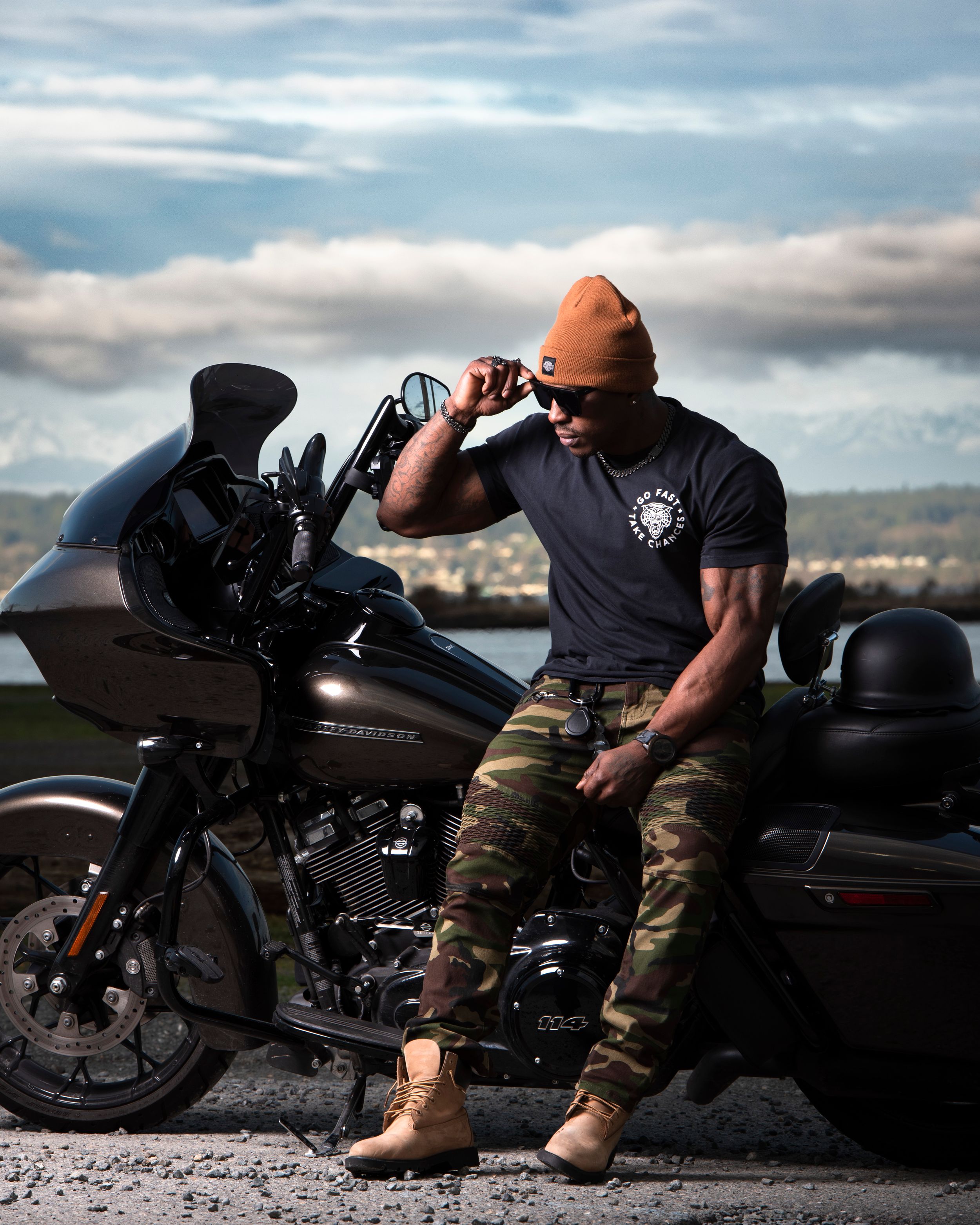 Motorcyclist in camo pants sitting on parked Harley Davidson against cloudy sky.