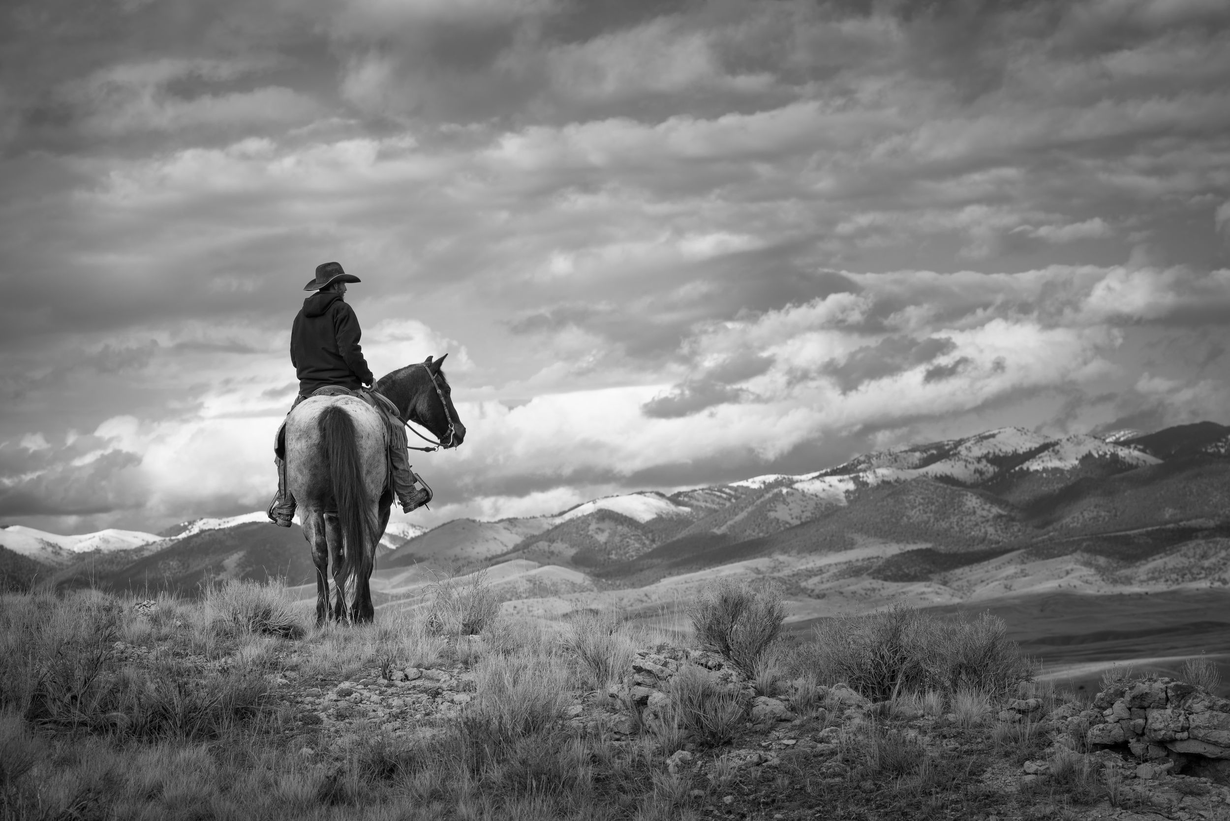 A cowboy rides horseback on a hilltop overlooking dramatic mountain landscapes in black and white photography.