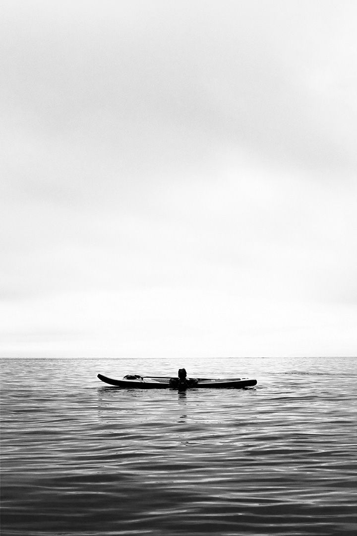 Black and white photo of a person lying on a paddleboard in calm ocean waters.
