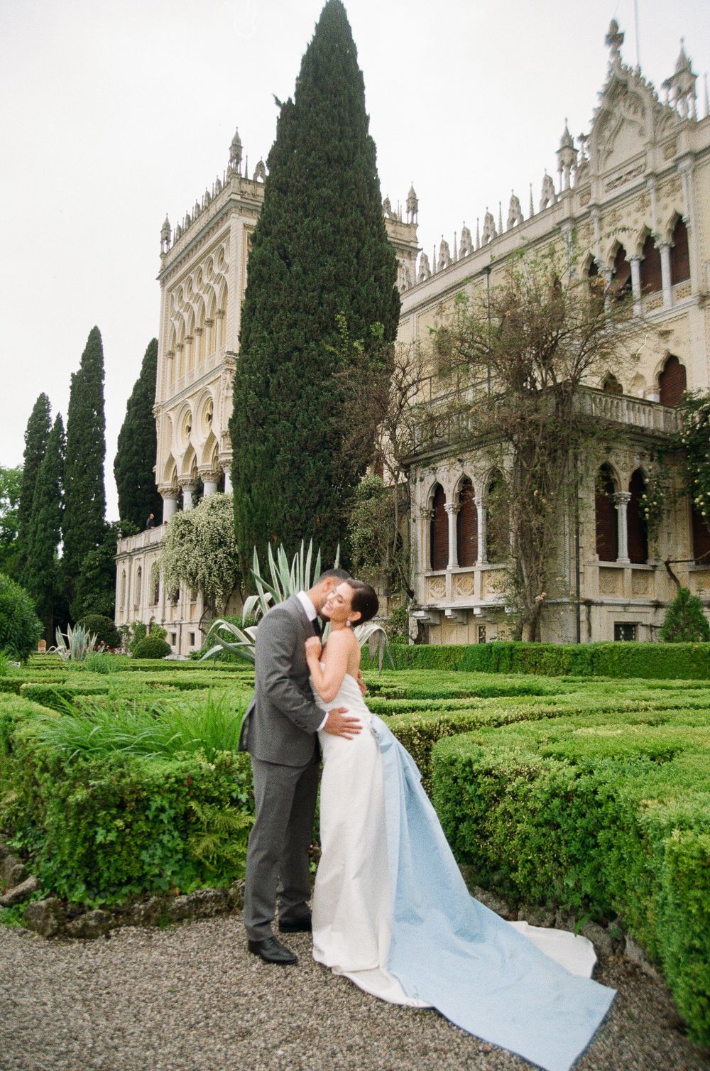 Romantic wedding photo at an ornate Italian villa with manicured gardens and cypress trees.