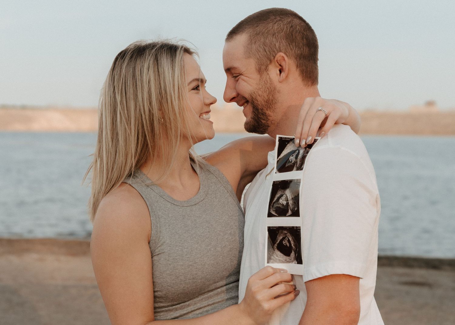 Couple sharing a tender moment while holding an ultrasound photo by a lake at sunset.