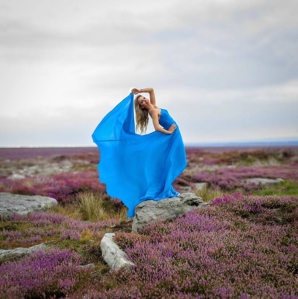 Figure in flowing blue fabric dress poses dramatically on purple heather moorland.