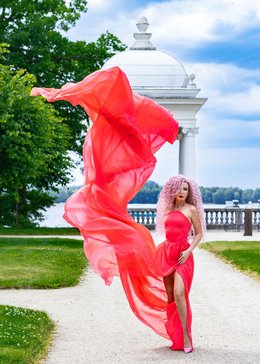 Flowing red dress billows dramatically against white architectural backdrop in garden setting.