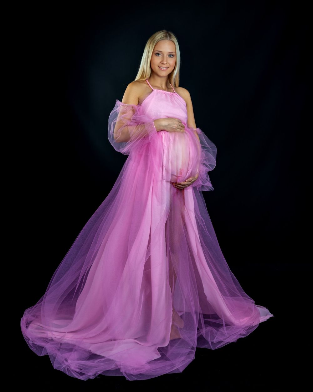 Maternity portrait in flowing pink tulle gown against dark background.
