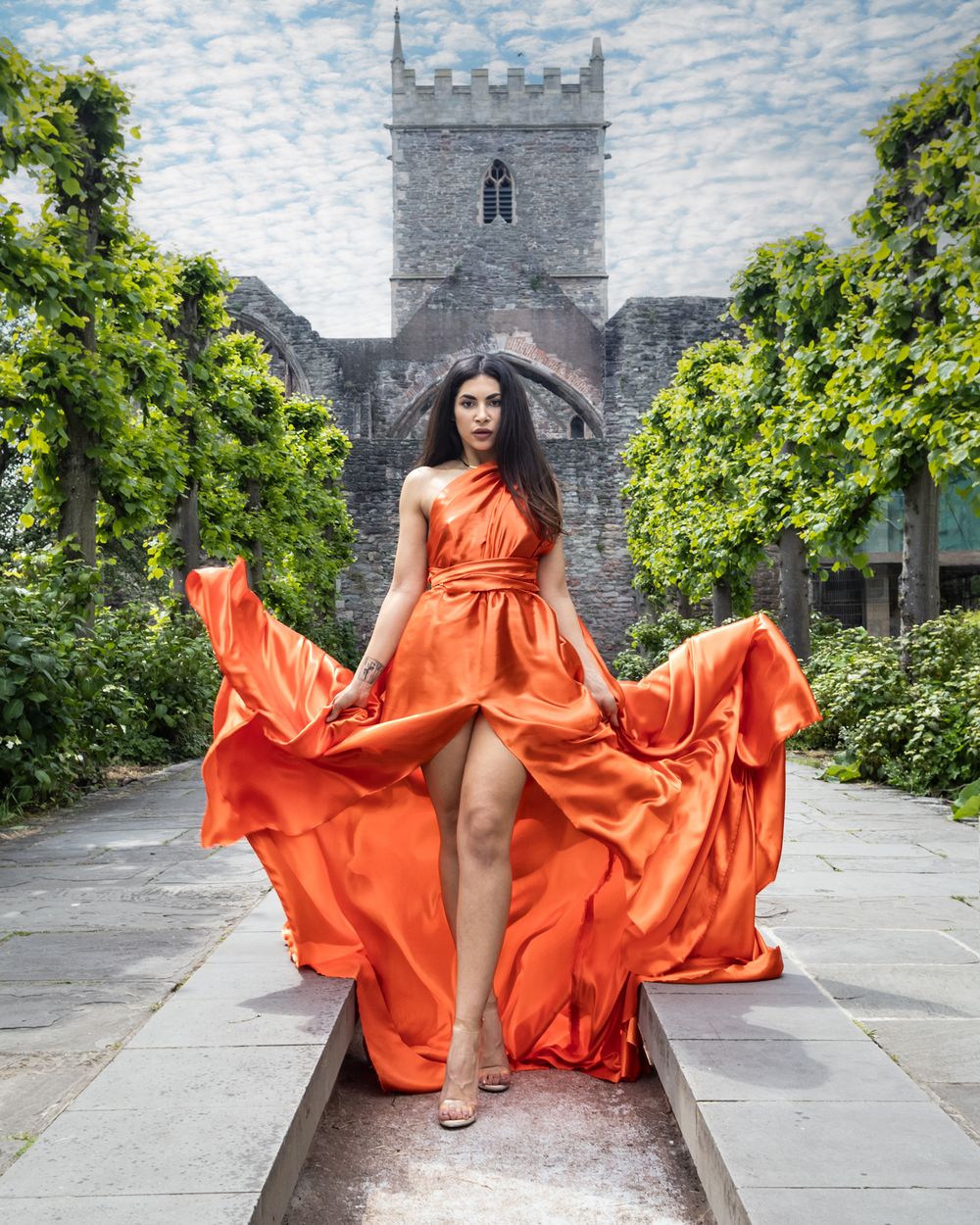 Flowing orange dress billowing dramatically in front of a stone church with archway.