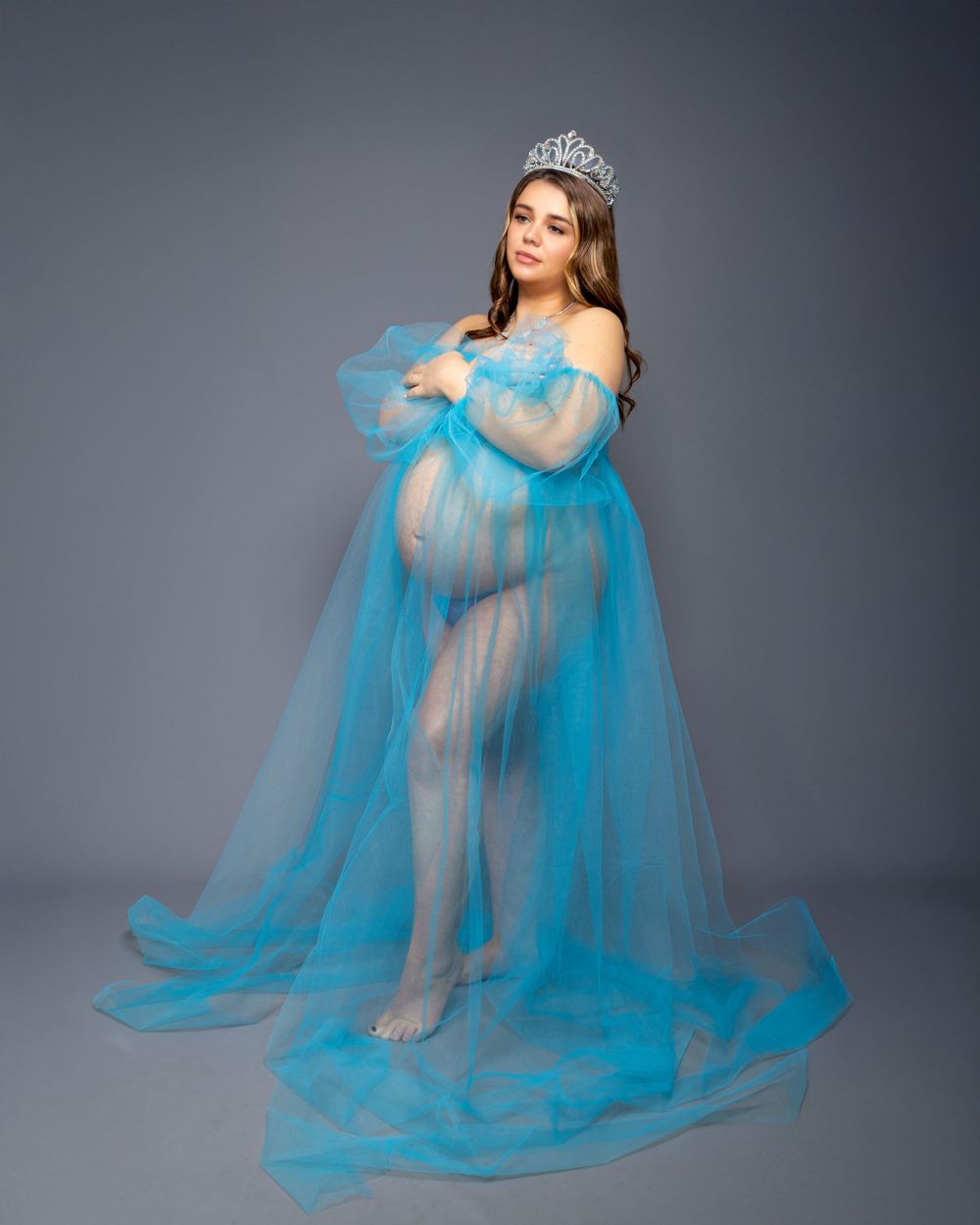 Elegant maternity portrait in sheer blue tulle gown and crystal tiara against grey background.