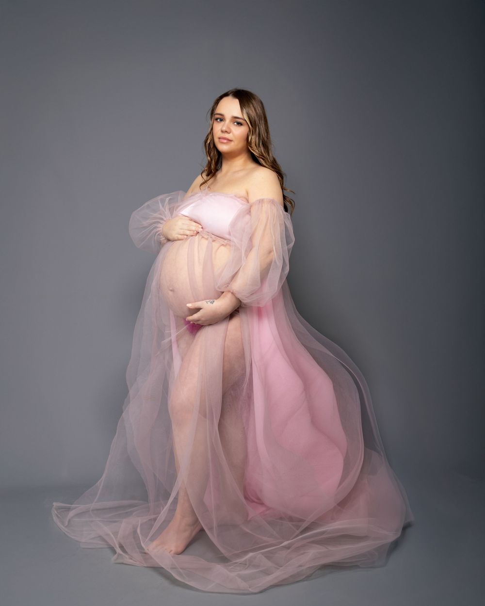 Dreamy maternity portrait in soft pink sheer gown against neutral grey background.