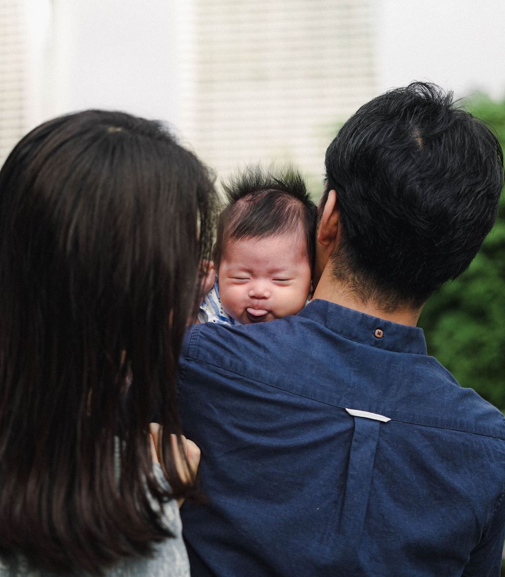 A laughing baby is held by two adults wearing casual clothing seen from behind outdoors.