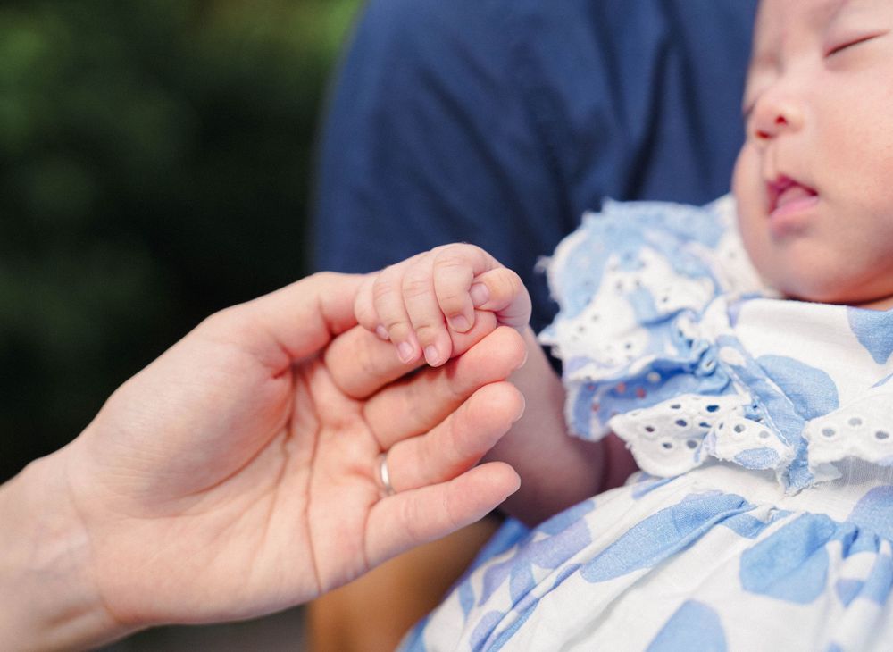 A tiny hand gently grasps an adult's finger while wearing a delicate blue and white lace outfit.