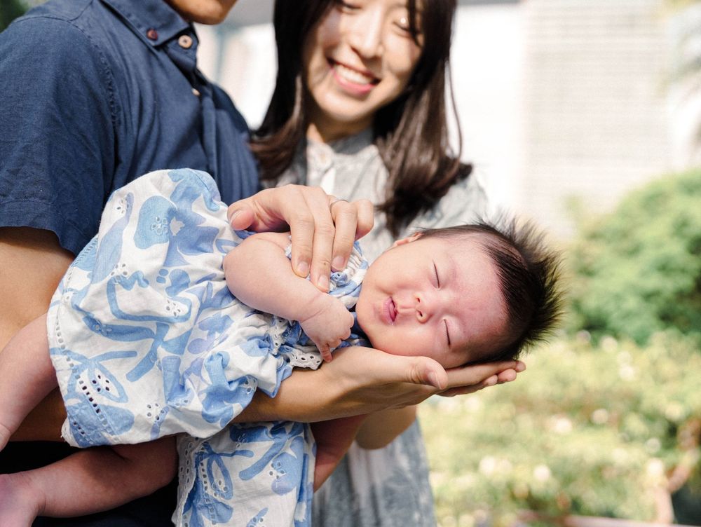 A peaceful sleeping baby wearing a light blue floral outfit is held tenderly outdoors on a sunny day.