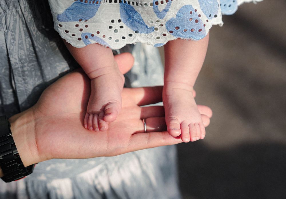 Tiny baby feet rest gently on an adult's hand while wearing a blue and white dress.