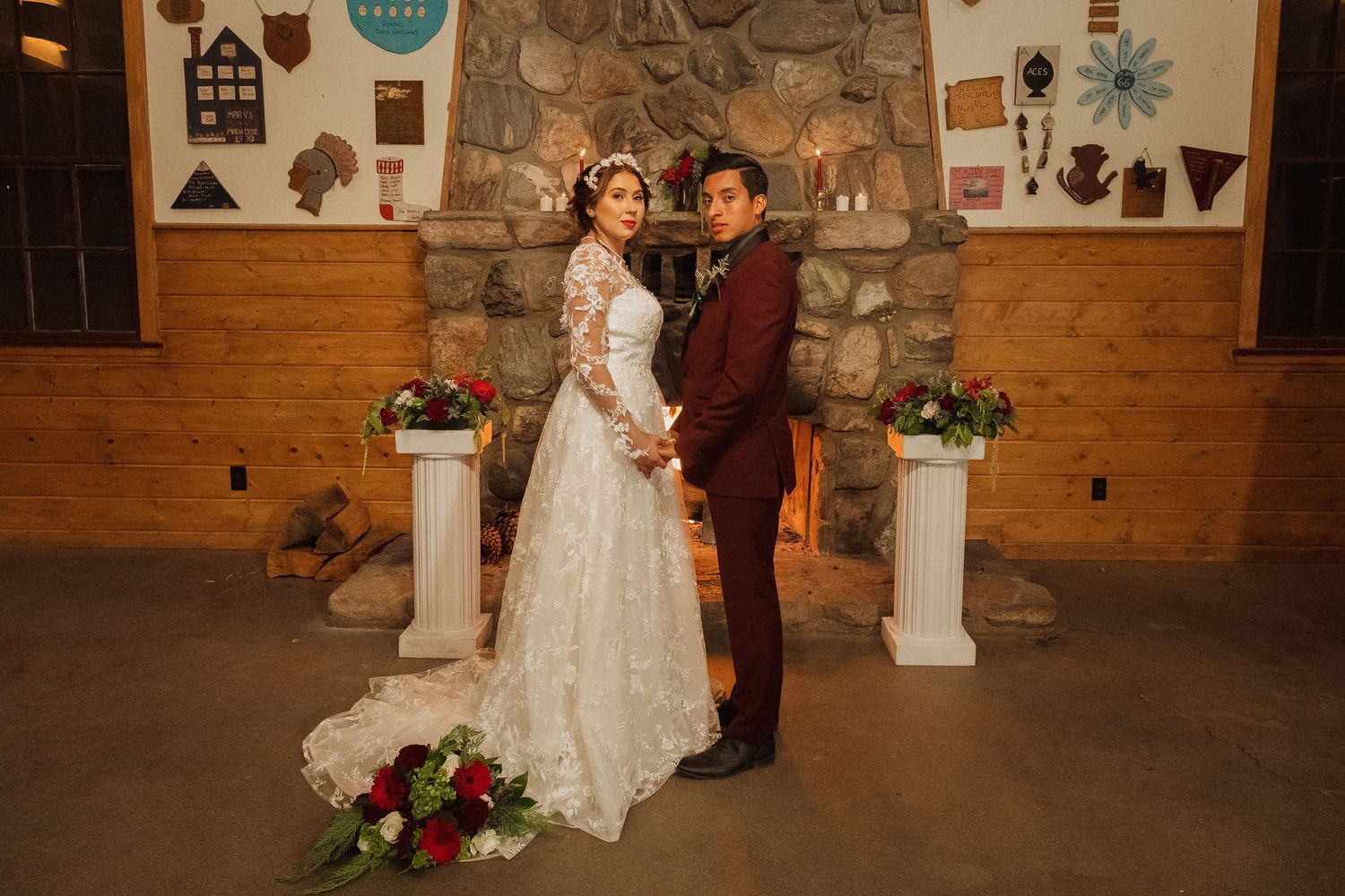 A couple poses in front of a stone and wood wall at their wedding with floral arrangements and decorative pedestals.
