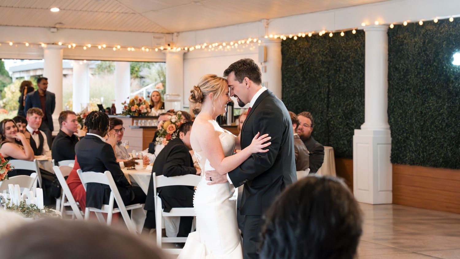 Couple dancing in elegant reception hall decorated with string lights and greenery wall.