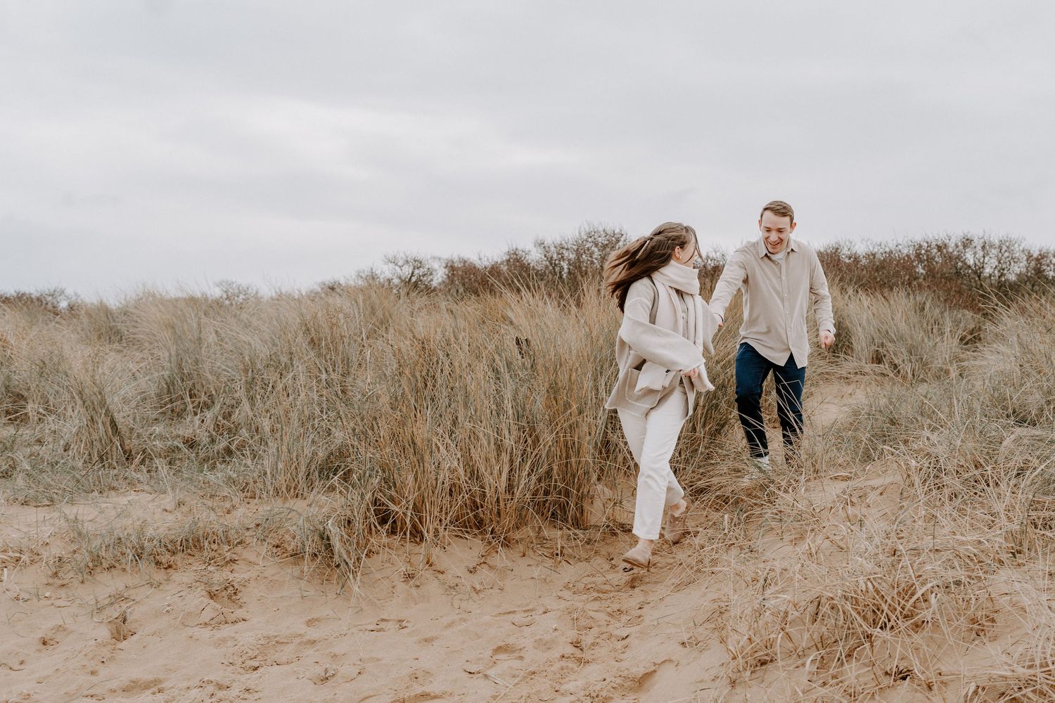 A couple are running down sand dunes on a beach. They are holding hands and the girl is looking back at her husband laughing.